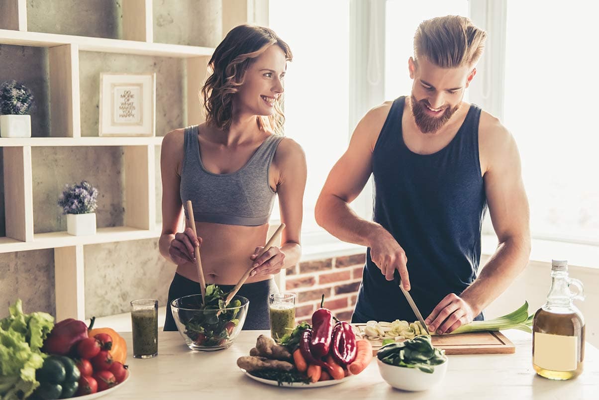 Pareja joven cocinando comida saludable: ensaladas, smoothies verdes y vegetales frescos